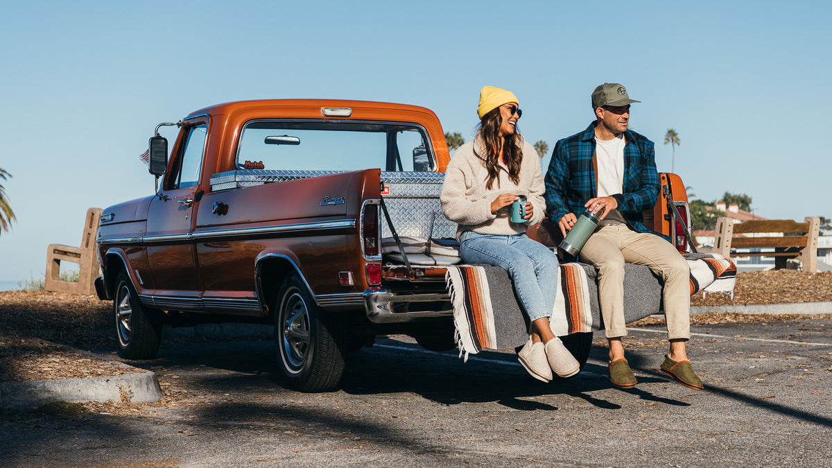 Couple relaxing on the tailgate of a vintage orange pickup with a striped blanket at the beach.