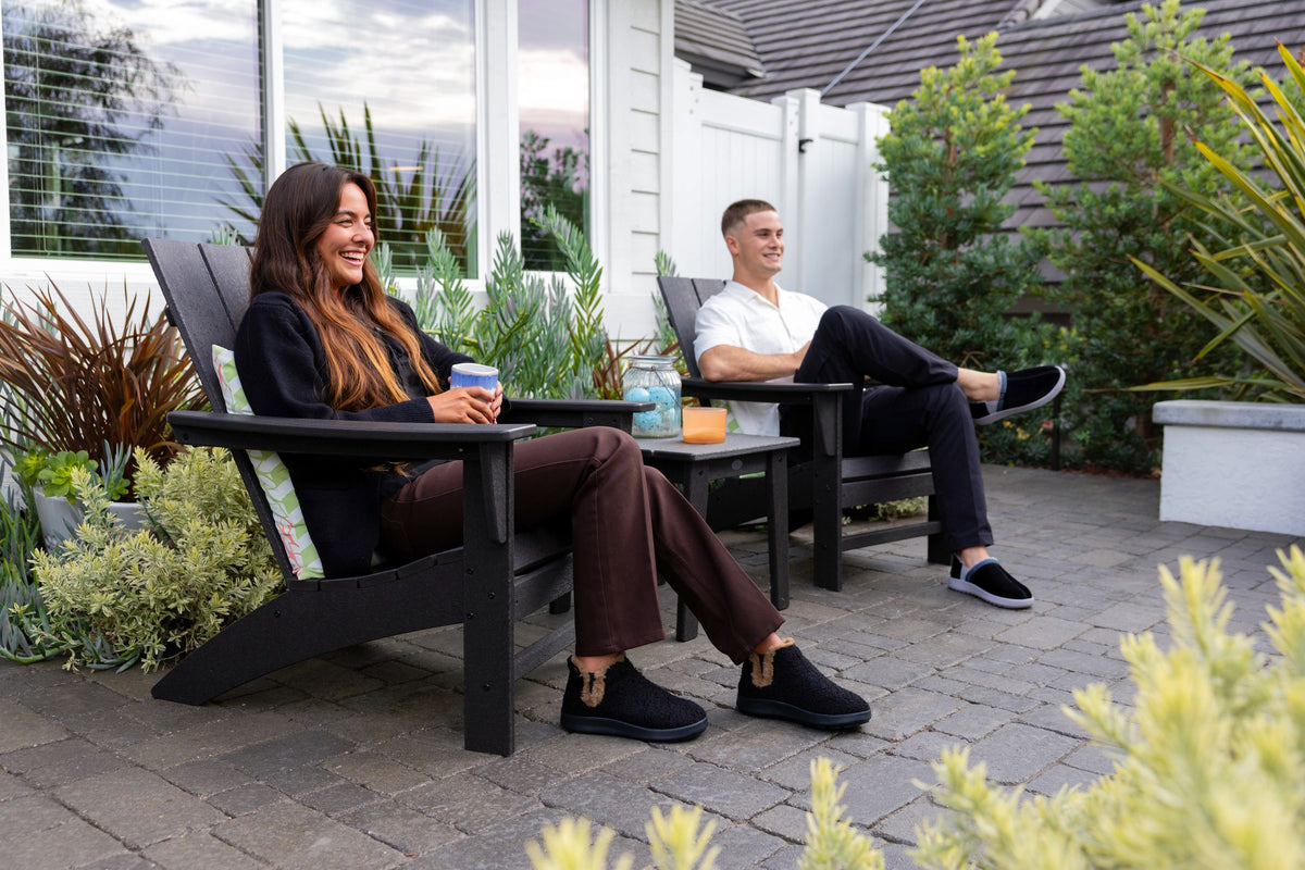 Two people relax in black outdoor Adirondack chairs on a paved patio, with a small table between them and plants and a house in the background.